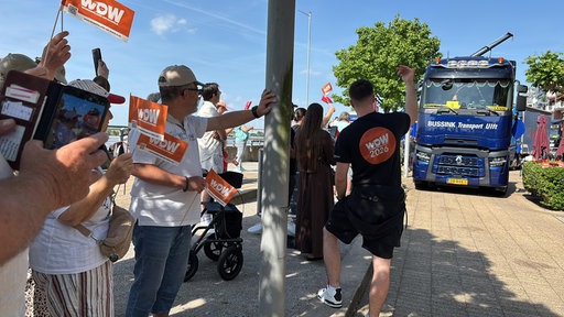 Mehrere Menschen stehen in Emmerich am Straßenrand und winken einem LKW-Konvoi