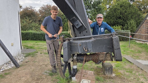 Zwei Männer stehen neben einemgroßen Holzbalken und lächeln in die Kamea.