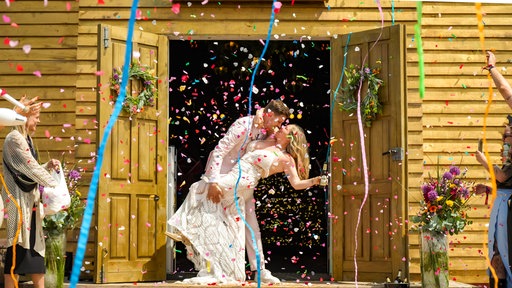 Ein sich küssendes Hochzeitspaar mit viel Konfetti vor einer Holzkirche auf einem Festival