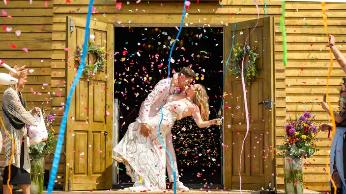 Ein sich küssendes Hochzeitspaar mit viel Konfetti vor einer Holzkirche auf einem Festival