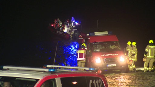 Einsatzkräfte stehen mit Einsatzwagen und einem Boot nachts am Rhein.
