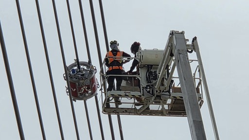 Menschen auf einem Kran areiten an einem runden Roboter an Drahtseilen einer Brücke.