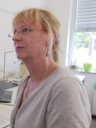 Eine Frau einem grauen Oberteil mit Brille steht vor ihrem Computer 