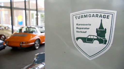 Auf einem Pfeiler ist ein weiß-grüner Sticker der Turmgarage und im Hintergrund steht ein organener Porsche