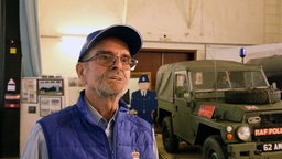 Ein älterer Mann mit Brille, Hemd, Weste und Mütze auf dem Kopf steht vir einem Militärfahrzeug