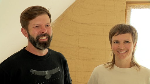 Ein Mann und eine Frau stehen auf einer Baustelle und lächeln in die Kamera.