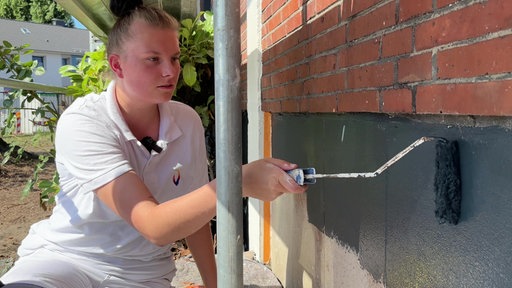 Eine junge Frau in weißer Malerkleidung streicht den unteren Teil einer Hausfassade