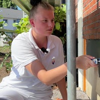 Eine junge Frau in weißer Malerkleidung streicht den unteren Teil einer Hausfassade