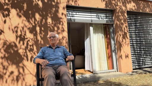 Ein älterer Mann mit Brille und weißen Haaren sitzt auf einem Stuhl vor einem Haus. Er lächelt in die Kamera.