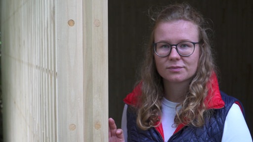 Landwirtin Hanna Bonsels trägt eine dunkelblaue Weste mit rotem Kragen. Sie steht neben einer Holzwand.