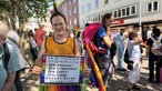 Marius-Felix-Aladini Femaro trägt ein orangenes TOp und ein Schild in seinen Händen.