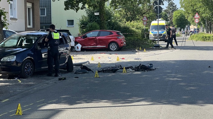 An einer Unfallstelle stehen drei stark beschädigte Autos. Auf dem Boden Einzelteile von Fahrrädern, ein Schuh und eine Schultasche, drumherum Polizeimarkierungen und Polizisten.