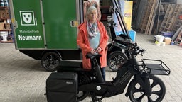 Birgit Pelc hat kurze weiße Haare und trägt einen roten Mantel. Sie steht vor dem Lastenwagen mit der Aufschrift "Malerbetrieb Neumann" und hinter einem schwarzen E-Bike.