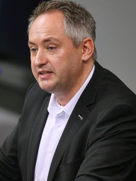 Ein Ein Mann mit kurzen dunklen Haaren. Er trägt ein schwarzes Sakko und ein weißes Hemd. Im Hintergrund ein blauer Stuhl.