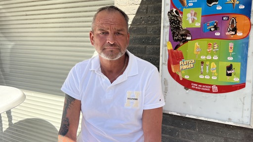 ein Mann in weißem T-Shirt sitzt vor einer Steinwand, in der rechten Bildecke ein Plakat mit Eiswerbung.