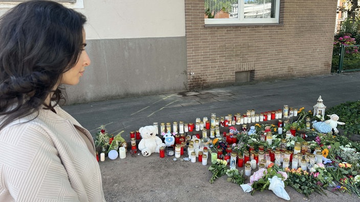 Eine dunkelhaarige Frau steht seitlich in der linken Bildecke und schaut auf eine Gedenkstätte mit Kerzen, Blumen und Stofftieren.
