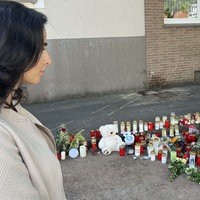 Eine dunkelhaarige Frau steht seitlich in der linken Bildecke und schaut auf eine Gedenkstätte mit Kerzen, Blumen und Stofftieren.