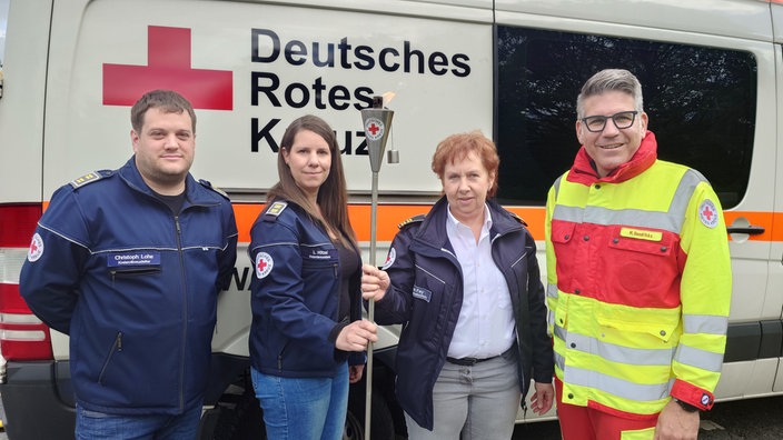 Vier Mitarbeiter des Deutschen Roten Kreuzes vor einem Auto des Deutschen Roten Kreuzes. Links und rechts stehen zwei Männer und in der Mitte zwei Frauen. Die beiden Frauen halten die Fackel zwischen sich.