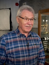 Walter Hoffacker hat ein blau-kariertes Hemd an, trägt eine Brille und hat graue, kurze Haare. Hinter ihm steht eine Vitrine mit vielen alten Kameras und Trophäen.