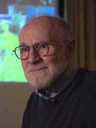 Werner Kraasch trägt einen blauen Pullover, hat eine schwarze Brille und einen weißen Bart. Hinter ihm ist eine Projektion an der Wand.