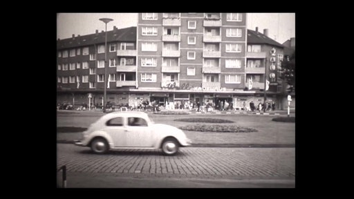 Eine schwarz-weiß Aufnahme eines weißen Autos, das auf einer Straße vor einem mehrstöckigen Haus fährt.