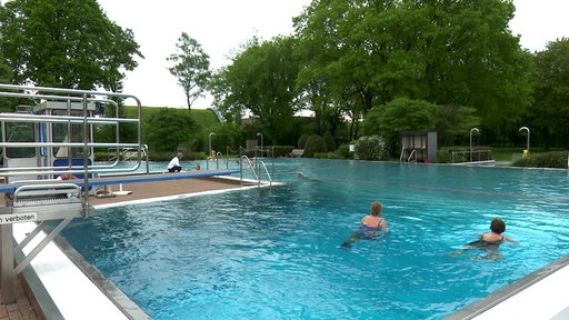 Zwei Frauen schwimmen in einem Schwimmbecken. Hinter dem Becken stehen Bäume und Büsche.