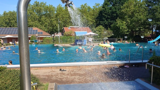 Viele Menschen sind im Schwimmbecken. Eine Dusche sprüht Wasser.