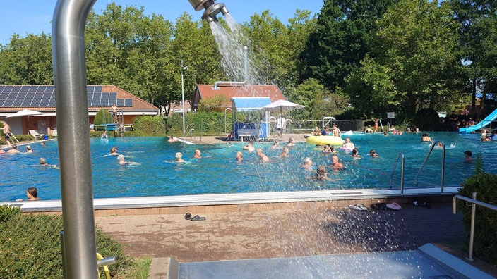 Viele Menschen sind im Schwimmbecken. Eine Dusche sprüht Wasser.