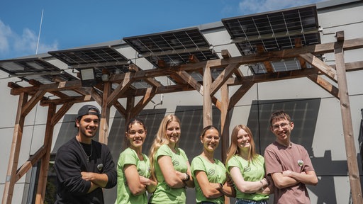 Jugendliche stehen mit verschränkten Armen vor einer Wand mit Balkenkonstruktion und Solarmodulen.