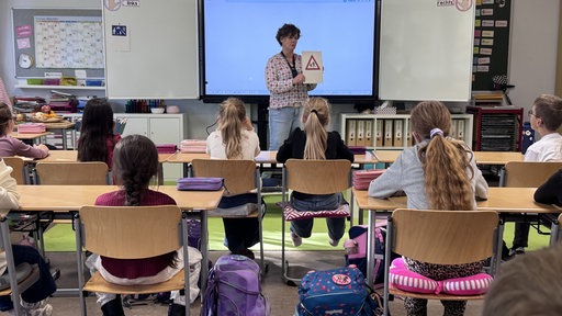 Kinder sitzen mit dem Rücken zur Kamera auf Stühlen und schauen zu einer Frau, die ein Papier in den Händen hält.