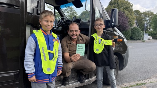 Ein Mann in brauner Uniform hockt vor einem Laster zwischen zwei Jungen mit neon-gelben Reflektoren. In der Hand hält er einen grünen Zettel mit der Aufschrift "Dankzettel"