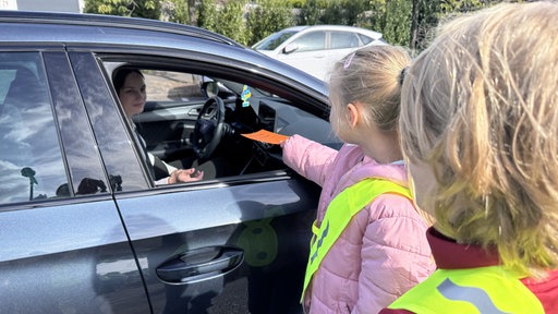 Zwei Mädchen geben einer Frau in einem Auto einen roten Zettel druch das geöffnete Fenster.