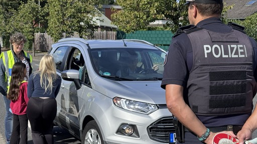 Drei Menschen stehen links an einem silbernen Auto. Davor sieht man den Rücken eines Polizisten.