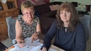 Zwei Frauen, Heidrun Reinders und Sabine Grundmann, blicken in die Kamera. Sie halten Dokumente in den Händen.