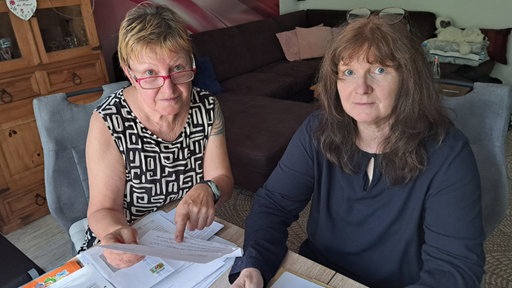 Zwei Frauen, Heidrun Reinders und Sabine Grundmann, blicken in die Kamera. Sie halten Dokumente in den Händen.