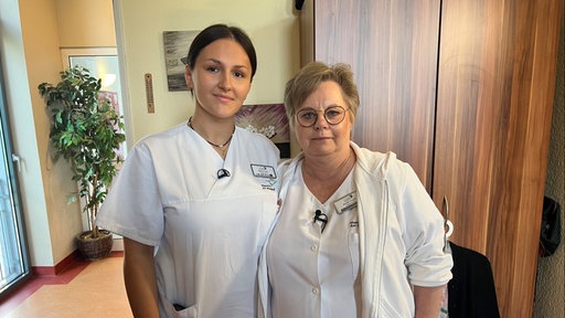 In Rheinberg arbeiten Melisa Trumic und Mariola Galwas in einem Pflegezentrum zusammen. Hier setzt man bewusst auf Teams aus jungen und älteren Pflegehilfskräften. Auf dem Bild haben sie beide weiße Pflegekleidung an und gucken in die Kamera.