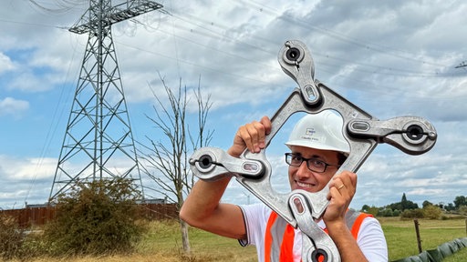 Ein Mann mit Warnweste hält ein Metallteil einer Stromleitung in der Hand. Im Hintergrund sieht man einen Strommasten und eine Leitung.
