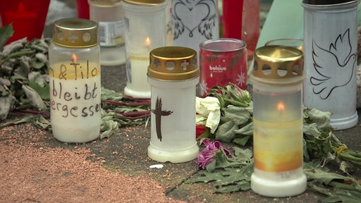 Auf einem Stück Asphalt stehenviele brennende Grabkerzen, dazwischen liegen Blumen