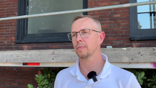 Ein Mann mit Brille, Bart und weißem Poloshirt steht vor einem Haus und schaut in die Kamera