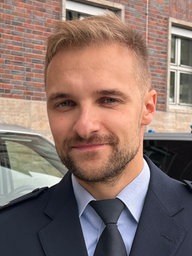 Ein junger Polizeibeamter in Uniform lächelt freundlich in die Kamera, hinter ihm sind zwei Polizeiautos im Anschnitt zu sehen