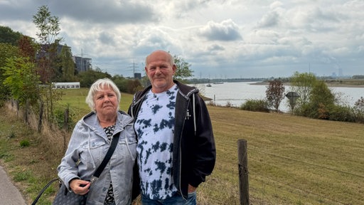 Ein älteres Ehepaar steht vor einer Wiese, im Hintergrund der Rhein
