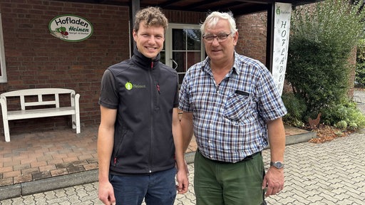 Julius Heinen und Peter Heinen stehen nebeneinander vor einem Haus und lächeln in die Kamera.