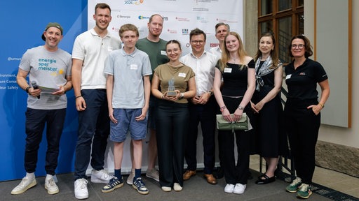 Eine Gruppe an Schülerinnen, Schülern und Erwachsenen steht bei einer Preisverleihung vor einer Werbewand. Das Mädchen in der Mitte hält eine silberne Trophäe in den Händen.