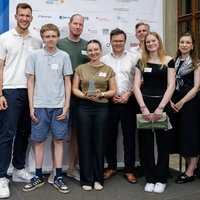 Eine Gruppe an Schülerinnen, Schülern und Erwachsenen steht bei einer Preisverleihung vor einer Werbewand. Das Mädchen in der Mitte hält eine silberne Trophäe in den Händen.