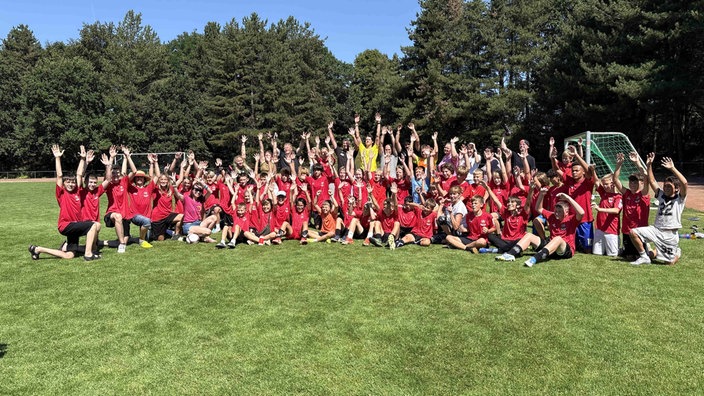 Ganz viele Kinder in roten Trikots sitzen auf einem Fußballfeld und werfen die Hände in die Höhe