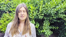 Frau mit langen Haaren steht vor grünem Busch