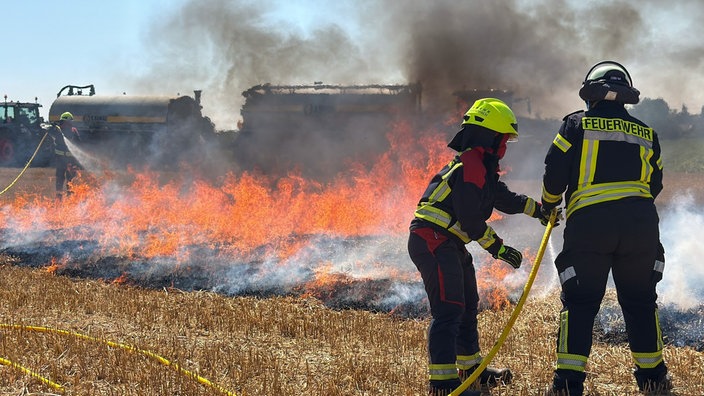 Feuerwehrleute stehen mit einem Wasserschlauch vor Flammen und versuchen, das Feuer zu löschen