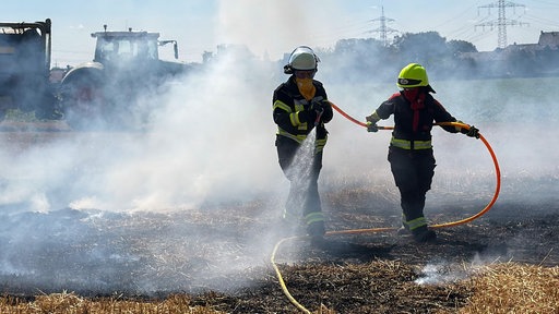 Zwei Feuerwehrleute stehen mit einem Wasserschlauch auf einem Feld, um sie herum verbranntes Stroh und dichter Rauch