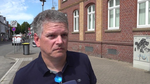 Ein Mann mit Polohemd steht ernst guckend auf einem Gehweg.