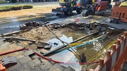 Asphalt ist aufgebrochen, man sieht Rohre in einer großen Wasserpfütze. Drumherum ist eine Baustellen-Absperrung. Ein Bagger und Arbeiter sind im Hintergrund zu sehen.